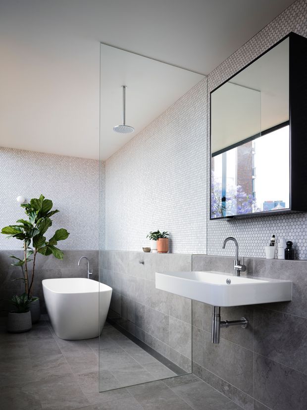 A contemporary bathroom with a freestanding white bathtub, wall-mounted white basin, large black-framed mirror, frameless glass shower screen, and chrome fixtures. Grey stone-look tiles cover the floor and lower walls, while small white mosaic tiles line the upper walls. A potted plant adds greenery, and natural light enters through the window.