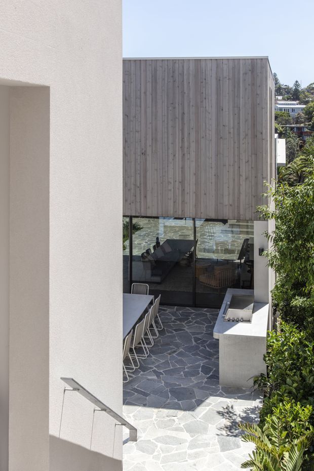 A contemporary multi-level residential courtyard viewed from an upper stair or landing. The space features a dark irregular flagstone-paved patio, a long dining table with chairs, a built-in outdoor kitchen or barbecue bench, floor-to-ceiling black-framed glazing, white rendered walls, and a tall vertical timber-clad upper volume. Lush planting borders the courtyard, with neighboring homes visible in the distance.