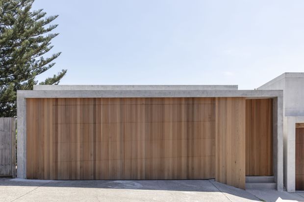 Front exterior view of a minimalist contemporary residence, dominated by a broad vertical timber-clad garage or entry door set within a smooth grey concrete facade. The flat roofline and crisp rectangular forms create a restrained modern composition, with a paved driveway in the foreground and a narrow adjacent timber door.