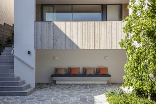 Exterior view of a contemporary multi-level home featuring a covered outdoor lounge beneath an upper balcony. The facade combines white rendered walls, vertical pale timber screening, dark-framed glazing, and irregular grey stone paving. A built-in bench with cushions sits beneath the balcony, beside a stairway and landscaped planting.