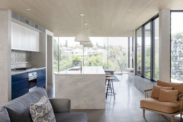 A bright open-plan kitchen and living area with a large marble-look island, deep navy lower cabinetry, integrated appliances, pendant lighting and floor-to-ceiling glazing. A grey sofa and tan armchair occupy the foreground, while leafy suburban views are visible through the windows. The space has a refined contemporary feel with a restrained neutral palette and strong indoor-outdoor connection.