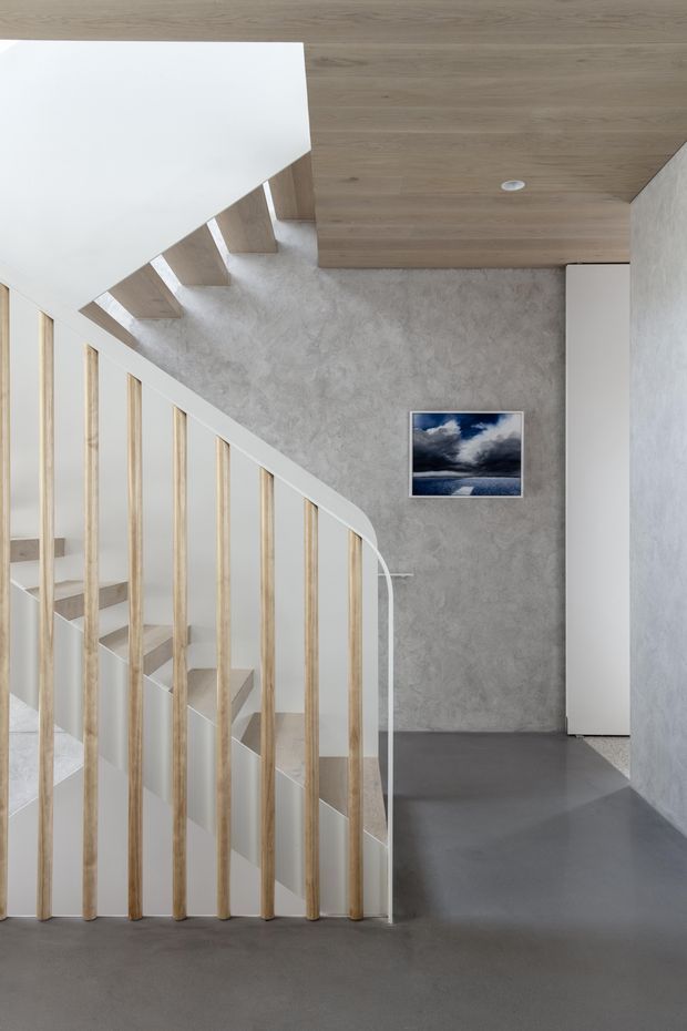 A minimalist interior stairway and landing. A sculptural white staircase with slim light-timber vertical balusters rises along a textured grey wall, beneath a warm timber-clad ceiling. The landing has a dark grey smooth floor, white built-in cabinetry or door, recessed lighting, and a small framed seascape artwork.