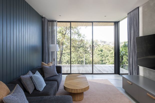 A contemporary living room with a dark vertical timber-panelled feature wall, deep charcoal sectional sofa, round sculptural timber coffee table, and a large wall-mounted television on built-in cabinetry. Full-height glazed sliding doors open to a timber deck, bringing in abundant natural light and views of greenery. Floor-to-ceiling curtains and a neutral rug soften the clean-lined space.