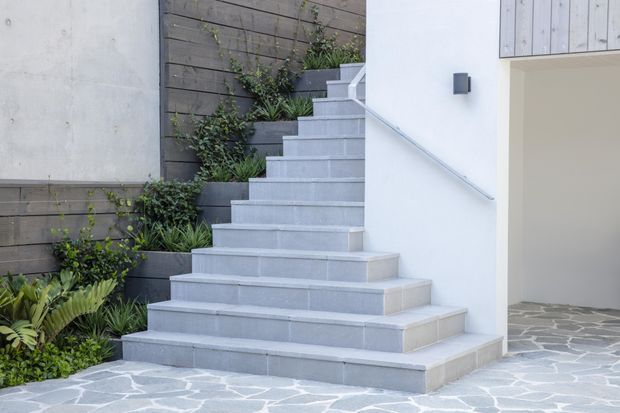 Exterior view of a modern entry stairway leading up alongside a white rendered building. Broad pale grey tiled or concrete steps have a simple metal handrail, with dark timber retaining walls and planted garden beds to the side. Irregular grey stone paving extends across the foreground and into a partially visible garage or entry opening.