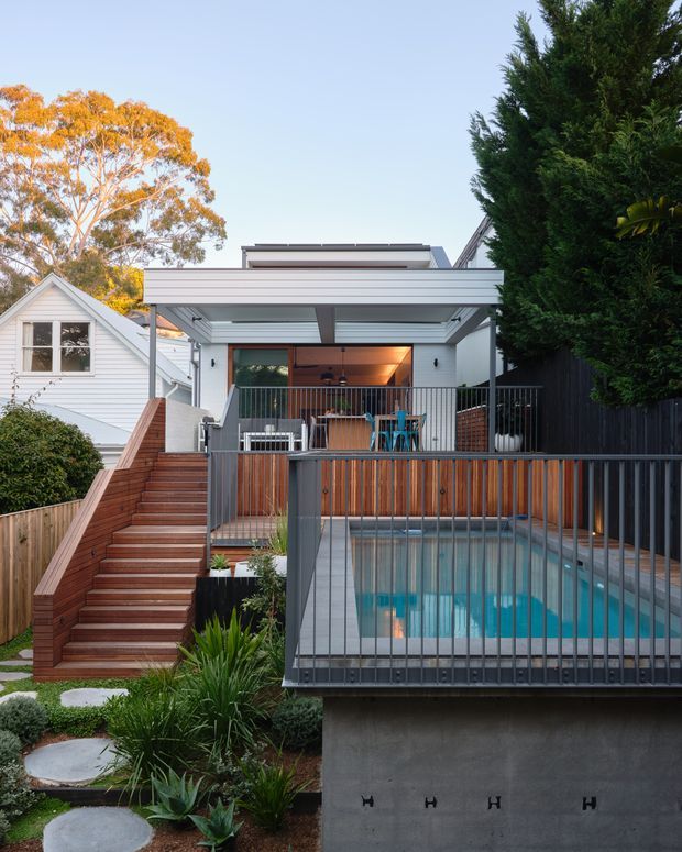 Exterior view of a contemporary multi-level home with a covered upper deck, outdoor dining area, timber stairway, landscaped garden, and a rectangular swimming pool enclosed by dark metal balustrades. The house combines white horizontal cladding, timber screening and decking, and concrete pool structure, with neighbouring residential fabric visible at the sides.