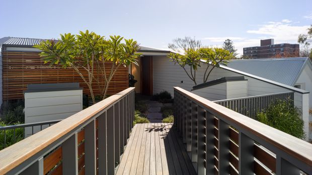 Contemporary residential exterior viewed from a timber pedestrian bridge or elevated deck, leading toward a low-profile home with corrugated metal roofing, horizontal timber screening, light cladding, and landscaped planting. Urban apartment buildings are visible in the distance.