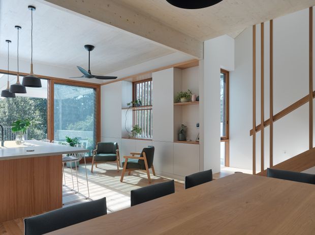 A bright, open-plan contemporary interior combining a kitchen, dining area and living space. The room features extensive light timber lining on the ceiling and cabinetry, a large timber dining table with dark upholstered chairs, a kitchen island, built-in shelving, vertical timber screening and large windows overlooking greenery. Natural light creates a warm, calm atmosphere.