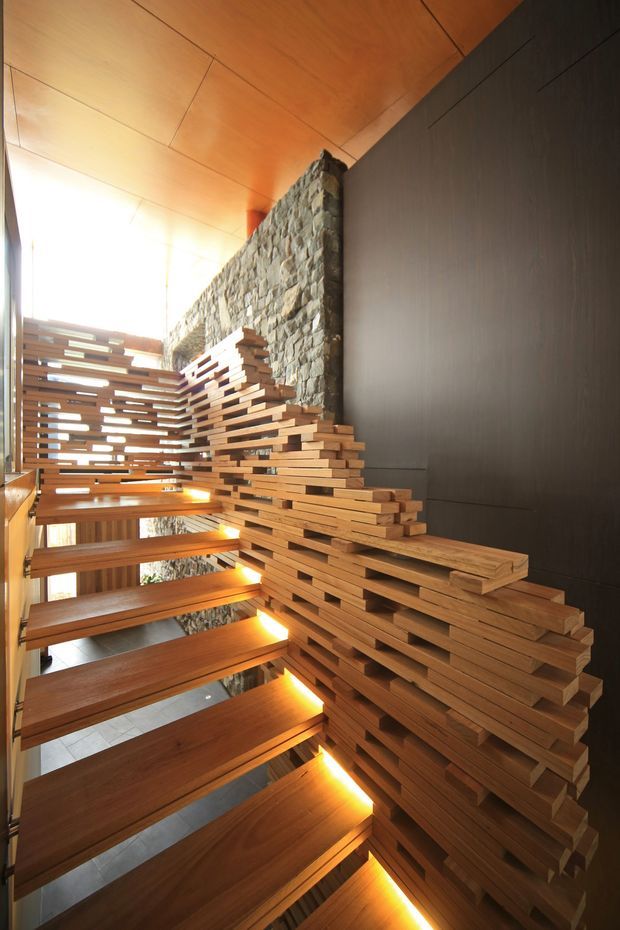 Architectural interior view of a dramatic open-riser timber stairway. Thick solid-wood treads and a sculptural stacked-timber balustrade rise alongside a rough natural-stone feature wall, with warm concealed lighting beneath the steps. The ceiling is clad in warm timber panels, while a dark smooth wall provides contrast. Bright daylight enters from the upper left, emphasizing the vertical void and layered construction.