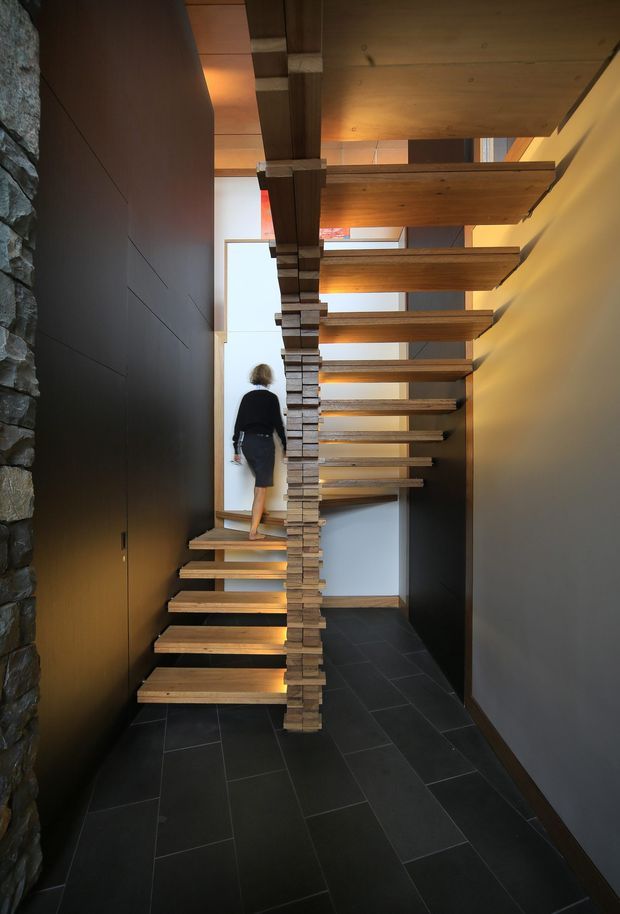 A dramatic interior stairway with floating timber treads arranged around a central stacked-stone structural spine. Dark tiled flooring, charcoal wall finishes, exposed timber ceiling elements, and stone wall cladding create a warm, high-contrast contemporary composition. A person is walking up the stairs, emphasizing the vertical scale and open void.