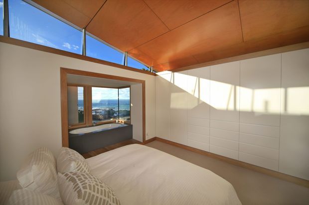 A bright contemporary bedroom with a low-profile bed, patterned neutral pillows, extensive white built-in wall cabinetry, and warm timber-lined ceiling and trim. High clerestory glazing and a projecting window seat or reading nook provide expansive ocean views, while strong afternoon sunlight creates geometric shadows across the walls.