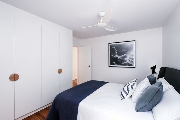 Bright, compact contemporary bedroom with a large upholstered bed, white bedding, navy throw, and layered blue-grey cushions. Full-height white built-in wardrobe doors with round timber pulls line the left wall, while a white ceiling fan, framed monochrome artwork, timber flooring, and a partially open door complete the space.