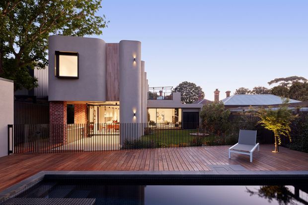 Contemporary residential exterior viewed from a timber deck beside a dark reflective swimming pool. The home features a sculptural two-storey form with smooth rounded grey plaster walls, a vertical timber-clad panel, exposed red brick at ground level, black metal fencing, and carefully integrated exterior lighting. A landscaped garden and neighbouring pitched-roof homes are visible beyond the property.