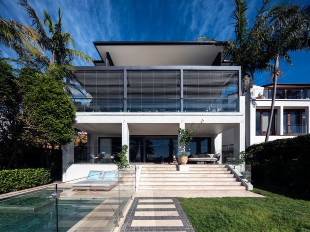 Contemporary multi-level residential facade with a broad flat roof, white rendered walls, extensive louvred upper-level glazing, and an open ground-floor terrace reached by wide pale stone steps. A paved path leads through a landscaped lawn to a glass-edged swimming pool, with tropical palms and hedging surrounding the property.