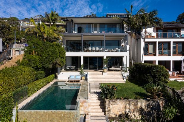 A multi-level contemporary coastal residence viewed from the garden, with extensive glazing, stacked balconies and terraces, a swimming pool, landscaped garden, stone retaining walls, and palm trees. The central home has a light neutral facade and dark-framed windows, with neighbouring residences visible on either side.