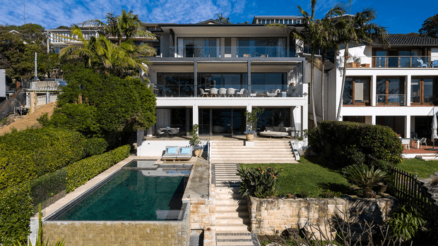 A large contemporary multi-storey residence on a landscaped sloping site, viewed from the rear garden. The home has extensive glazed openings, broad terraces and balconies, pale rendered and stone-clad surfaces, and a long rectangular swimming pool with stepped access and manicured planting.
