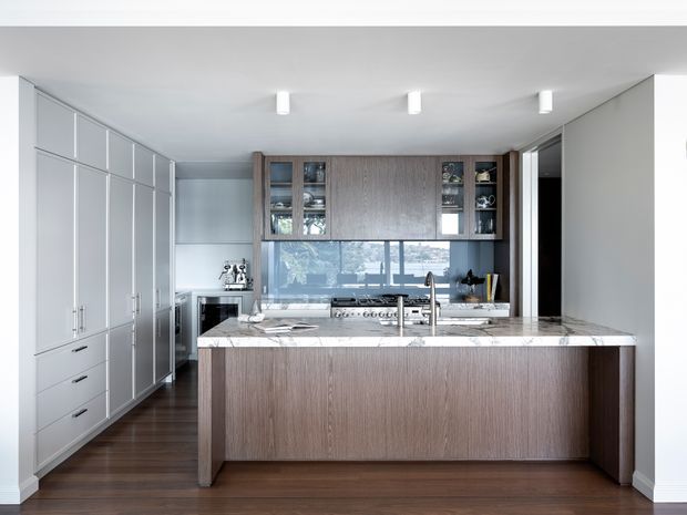 Contemporary kitchen with a large central island, integrated white cabinetry along the left wall, dark timber veneer island and overhead joinery, white marble-look benchtops, stainless steel appliances, and a wide window above the rear counter. The space is clean, bright, and highly tailored, with dark timber flooring and recessed linear lighting.