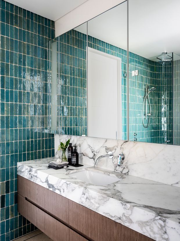 A refined contemporary bathroom vanity with a large white marble benchtop and integrated basin, warm wood-grain drawer cabinetry, and a wall-to-wall mirrored cabinet. The walls are clad in glossy deep teal rectangular tiles with contrasting grout. A frameless glass shower enclosure with overhead and handheld fixtures is visible in the reflection.