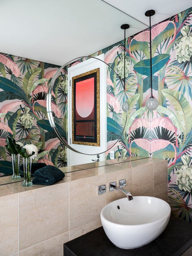 A richly styled bathroom vanity with a round vessel basin set on a dark benchtop, pale stone wall tiling, chrome wall-mounted tapware, and a large circular mirror. The walls feature bold tropical botanical wallpaper in pink, green, teal, and cream, complemented by black pendant lights and framed artwork.
