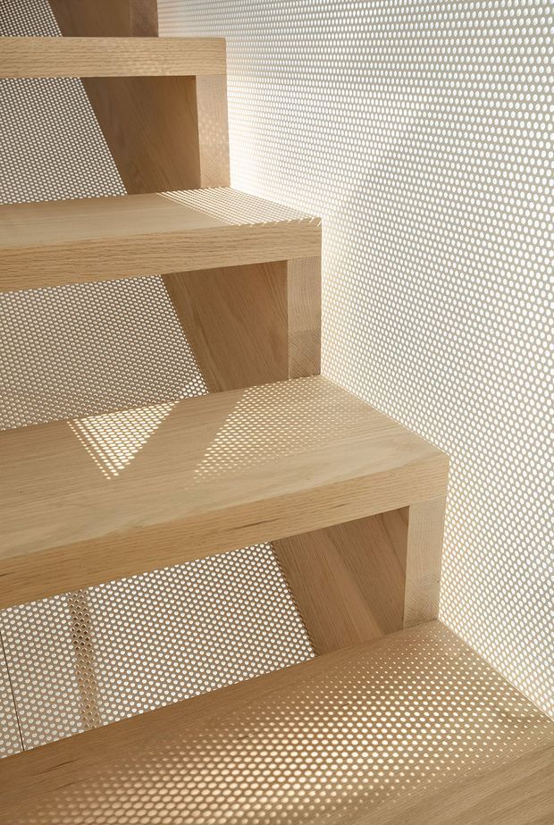 Close-up architectural detail of a light oak timber staircase enclosed by a perforated metal screen. Sunlight passes through the circular perforations, casting a strong dotted shadow pattern across the treads and risers. The image emphasizes warm natural timber, geometric repetition, and refined minimalist construction.