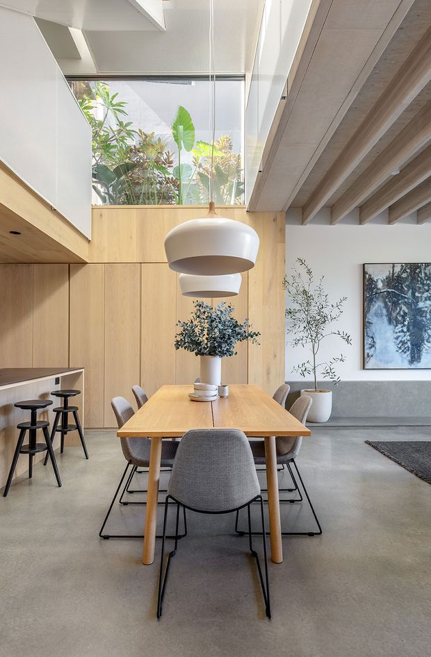 Bright contemporary dining area with a large timber dining table and grey upholstered chairs, set beneath oversized white pendant lights. The room features warm timber wall cabinetry, exposed timber ceiling beams, polished concrete flooring, a compact kitchen or bar zone with stools, indoor planting, and a glazed skylight bringing in abundant natural light.