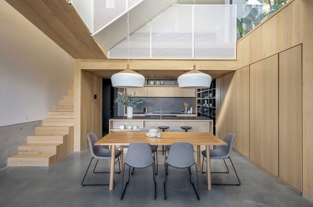 A bright, open-plan contemporary kitchen and dining area beneath a mezzanine. Pale timber wall panelling and cabinetry frame a dark kitchen with a stone island, while a timber dining table sits below two large pendant lights. A floating timber stair rises along the left wall, and a glazed balustrade encloses the upper level. The space has a polished concrete floor and a restrained palette of timber, grey, white and black.