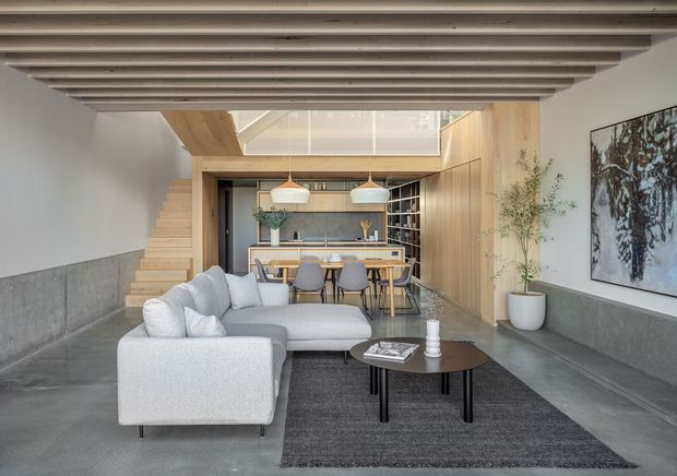 A contemporary open-plan living and dining area with a light grey sectional sofa, dark round coffee table, and textured charcoal rug in the foreground. The space features a dramatic exposed timber-beam ceiling, polished concrete flooring, a broad concrete wall ledge, warm timber cabinetry and wall panelling, a compact kitchen with island dining, pendant lights, open shelving, and a skylit double-height upper zone. A timber staircase rises along the left side, while artwork and potted greenery add restrained decorative elements.
