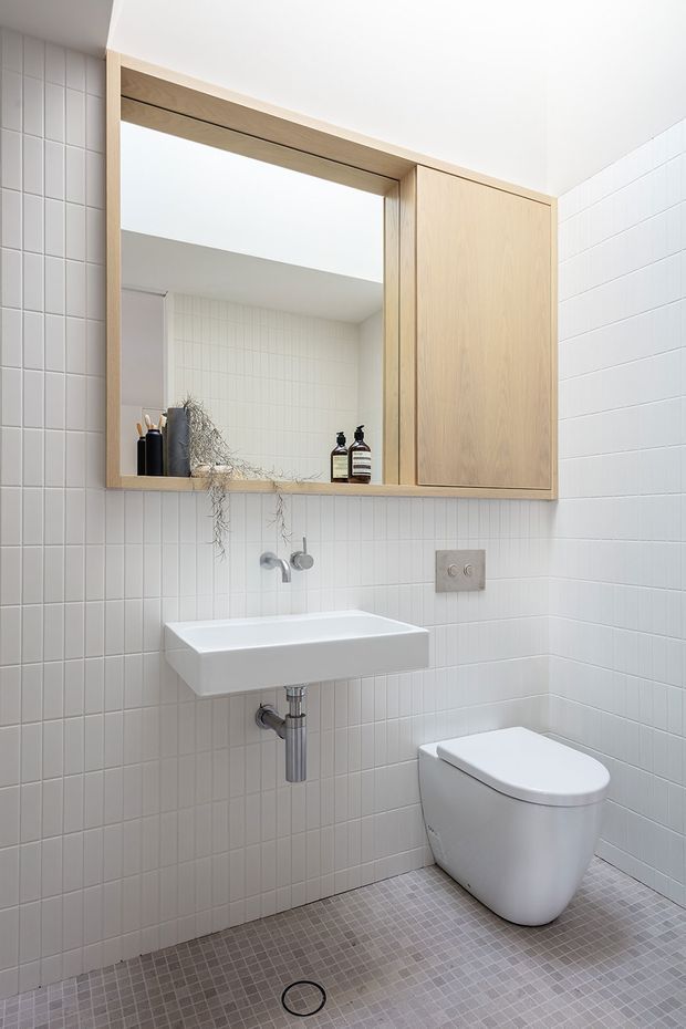 Bright compact bathroom featuring full-height white square wall tiles, small grey mosaic floor tiles, a wall-mounted white basin with exposed chrome plumbing, a streamlined white toilet, and a large light timber mirrored cabinet with open shelving and integrated storage. The space has a clean, minimalist fit-out with subtle natural styling from small plants and toiletries.