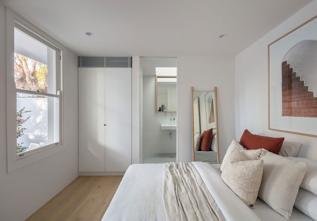 Bright, compact contemporary bedroom with a white upholstered bed, layered white bedding, and rust-red accent cushions. The room has pale timber flooring, white walls, built-in white wardrobes, a full-length mirror, a small adjoining bathroom visible through the opening, and a framed architectural artwork. Natural light enters through a large window, creating a clean and airy atmosphere.
