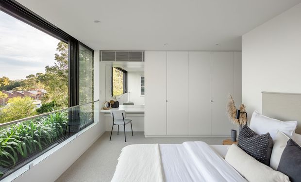 Bright, refined bedroom with a large corner of floor-to-ceiling black-framed glazing overlooking greenery and neighbouring homes. The room features a low-profile upholstered bed with white bedding and dark accent cushions, pale carpet, and a full wall of smooth white built-in wardrobes incorporating a compact desk and chair. The overall design is clean, contemporary, and uncluttered.