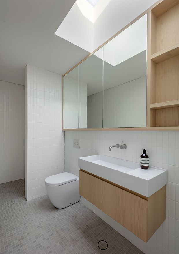Bright, minimalist bathroom with a wall-mounted timber vanity, integrated white rectangular basin, wall-mounted tapware, mirrored overhead cabinetry, open timber shelving, white tiled walls, and small grey floor tiles. A skylight brings natural light into the space, while a concealed toilet sits beside the tiled shower area.