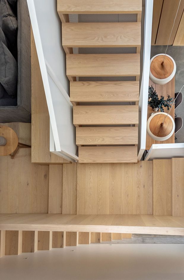 Top-down view of a contemporary timber staircase and landing. The stair treads, landing floor and adjacent built-in elements are finished in light natural wood, with white painted side walls and a perforated dark metal or mesh backing between open treads. A partial living area with a dark sofa and small round timber table is visible beside the stairs, along with decorative circular timber objects and greenery.