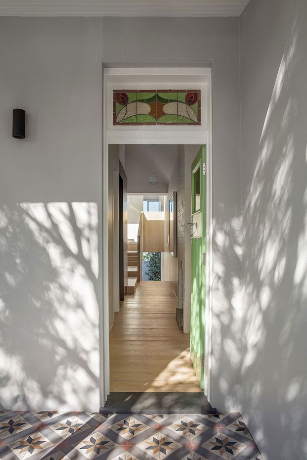 A bright residential entryway looking through an open doorway into a narrow timber-floored hallway. The foreground has patterned geometric encaustic-style floor tiles, white walls and trim, a green glazed door, and a decorative stained-glass transom panel with a floral and geometric motif. Sunlight casts leafy shadows across the walls, while timber stairs, built-in joinery, and glimpses of planted greenery are visible deeper inside.