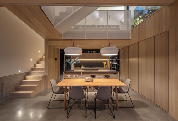 Warm contemporary open-plan dining and kitchen area with a substantial timber dining table, upholstered chairs, pendant lights, and integrated timber cabinetry. A floating stair rises along the left wall to a mezzanine or upper landing enclosed with clear glass balustrading. The space features polished concrete flooring, extensive timber wall and ceiling lining, concealed kitchen joinery, and soft integrated lighting.