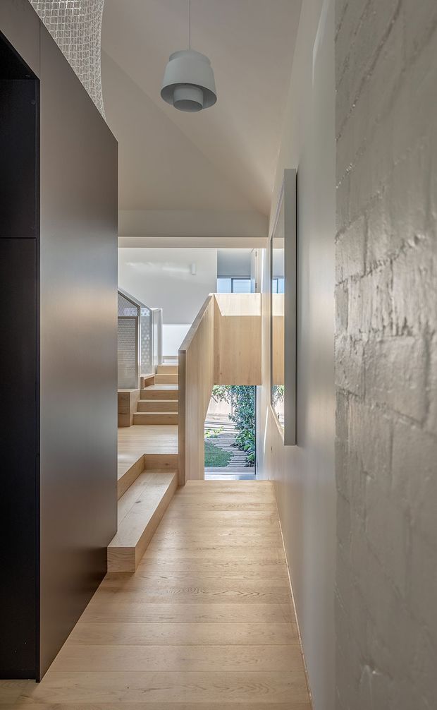 A narrow, light-filled interior circulation space with pale timber flooring and stepped stairs leading toward a glazed opening. Dark built-in joinery lines the left side, while a textured white masonry wall runs along the right. Pale timber balustrade and partition elements create layered levels, with a sculptural pendant light overhead and glimpses of greenery outside. The design is minimalist and contemporary, with a restrained neutral palette and strong linear geometry.