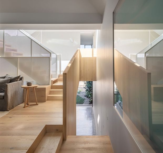 Bright contemporary split-level interior centred on a timber staircase and landing. Pale timber steps, flooring, and balustrade panels frame a partially visible living area with a sofa on the left. Translucent patterned screens, white walls, high-level glazing, and a large reflective glass panel create a light-filled, layered circulation space with views toward an exterior garden path.