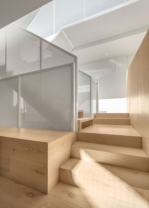 Bright minimalist interior stairway with broad pale timber steps and integrated timber storage or platform joinery. A perforated white metal balustrade and partition screens the stair opening, while white acoustic-looking ceiling panels and clerestory-style glazing create an airy, double-height feel. Strong natural light falls across the stairs and timber surfaces.