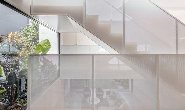 A bright, minimalist interior view focused on a white stairway and upper landing. A perforated or mesh-style white balustrade screens the lower level, while large floor-to-ceiling glazing on the left overlooks lush planting. White walls, ceiling planes, and stairs create a clean, airy architectural composition, with a partially visible living area below.