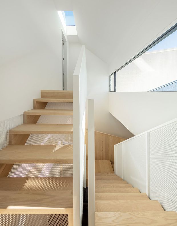 Bright minimalist interior view of a split stairway with two flights of pale timber stairs, white walls, perforated metal balustrade panels, and a high horizontal window bringing in daylight. A narrow skylight above the upper landing adds natural light, while the stair geometry and void create a sculptural architectural composition.