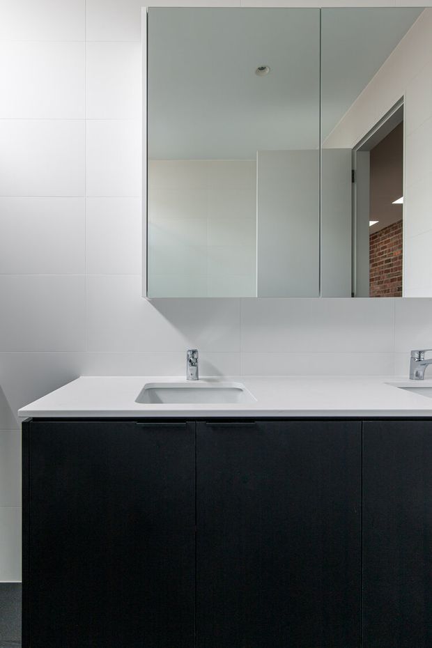Modern minimalist bathroom vanity with a large frameless mirrored cabinet, white tiled walls, integrated white basin and benchtop, chrome tapware, and dark flat-panel cabinetry. An open doorway reveals a brick-lined adjoining space.