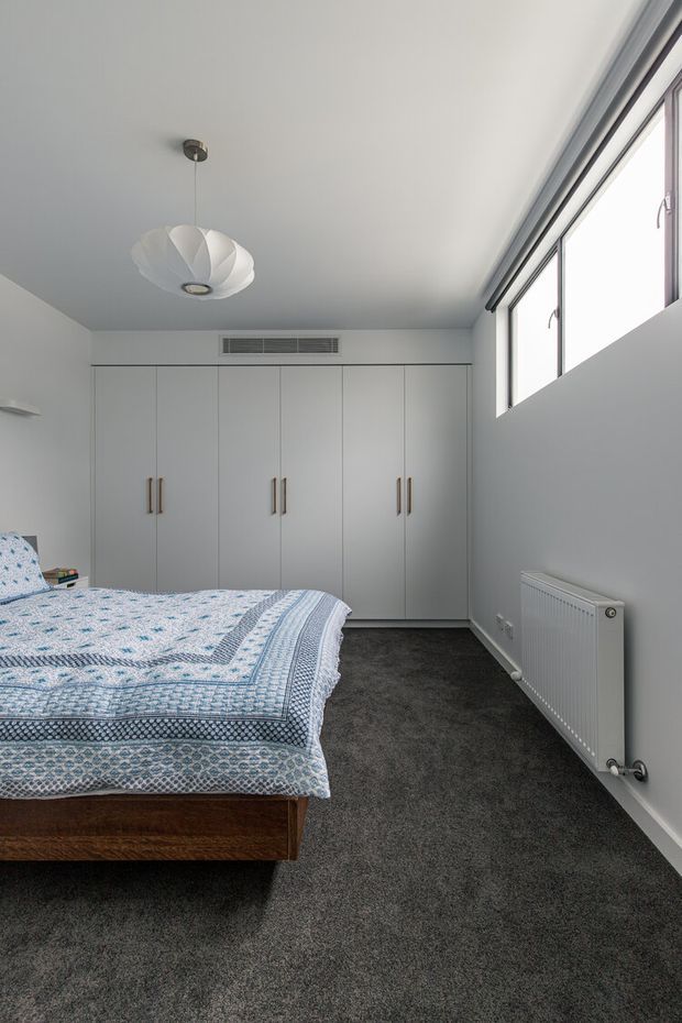 Bright, minimalist bedroom with a large upholstered or timber bed partly visible in the foreground, patterned blue-and-white bedding, full-height white built-in wardrobes with slim timber handles, dark grey carpet, white walls, a sculptural pendant light, and high horizontal windows providing abundant natural light. A wall-mounted radiator sits beneath the windows.