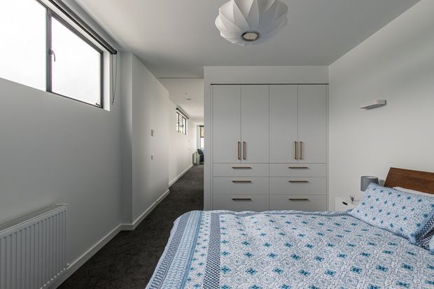 A bright, compact bedroom with a double bed in the foreground, patterned blue-and-white bedding, a timber bedhead, built-in white wardrobes and drawers, dark carpet, and white walls. A narrow hallway with windows and a radiator extends beside the bedroom, while a sculptural white ceiling light provides a decorative focal point.