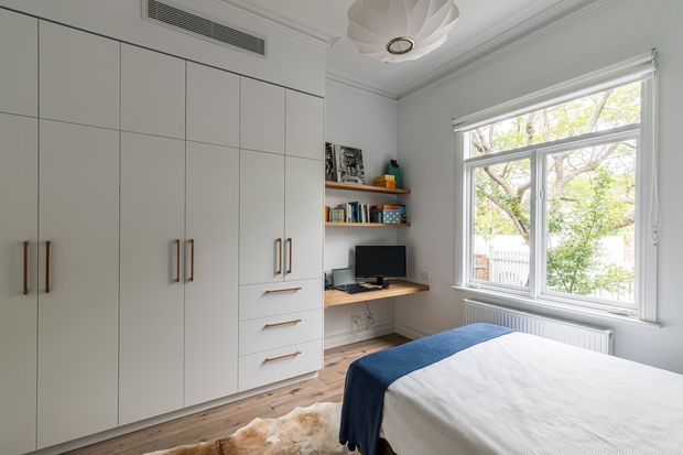 Bright, compact bedroom with integrated floor-to-ceiling white wardrobe cabinetry, a small built-in desk and open shelving, a large traditional sash-style window, timber flooring, and a neatly made bed with blue and white bedding. A sculptural pendant light and ornate ceiling detailing add a refined character.