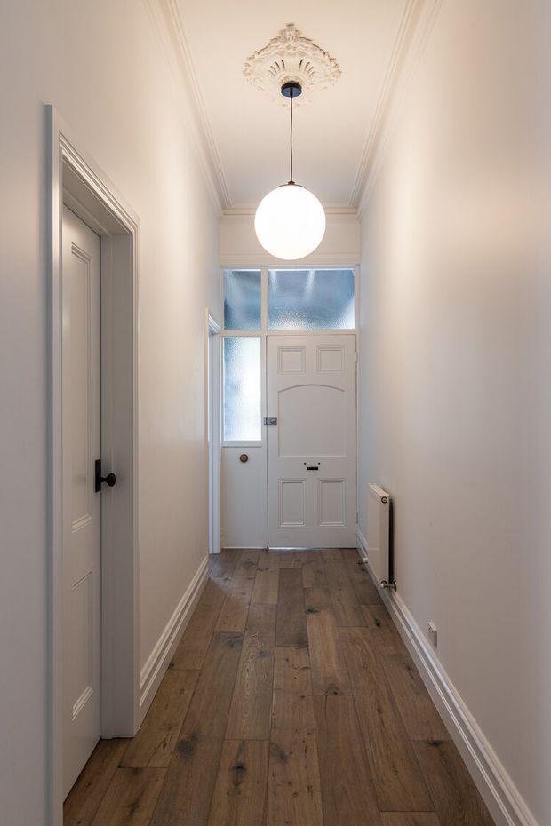 A narrow, refined residential hallway leading to a paneled front door with frosted glass sidelights and transom windows. The space features warm timber plank flooring, pale painted walls, detailed white cornices and skirting, an ornate ceiling medallion, and a simple globe pendant light. A paneled interior door and radiator line the corridor.