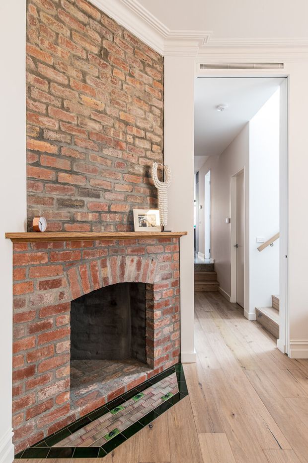 Interior view of a traditional hallway or entry passage featuring a prominent exposed red-brick fireplace with a timber mantel and decorative glazed tile hearth border. Pale painted walls, ornate white cornices, light timber flooring, and a staircase and corridor extend into the background.