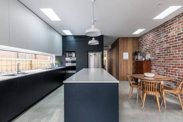 A contemporary open-plan kitchen and dining area with a long dark island, matte black cabinetry, pale stone benchtops, integrated appliances, polished concrete flooring, exposed red brick walls, and warm vertical timber slat detailing. Large windows and roof skylights provide abundant natural light, while a timber dining table and mid-century-inspired chairs sit beside a dark timber sideboard.