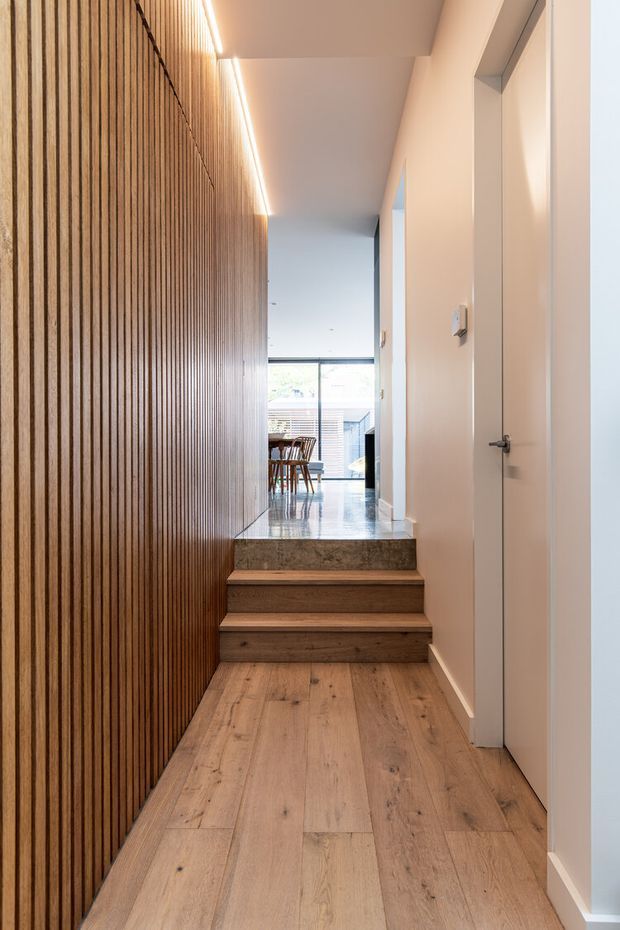 A narrow, contemporary interior hallway leading up a short set of timber steps toward a bright dining area. The hallway features warm wide-plank timber flooring, a full-height vertical timber slat feature wall on the left, white plaster walls and doors on the right, recessed doorways, and linear integrated LED lighting along the ceiling. A polished stone or marble-clad stair landing creates a contrasting finish.