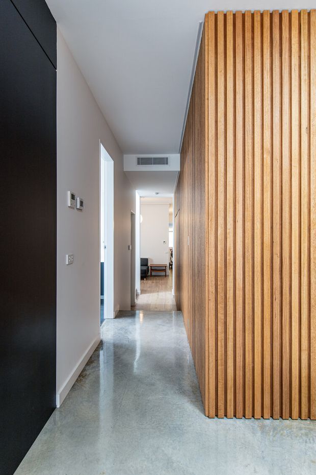 A narrow contemporary hallway with a polished grey concrete floor, pale painted walls, and a prominent full-height vertical timber slat feature wall or screen. A dark charcoal built-in surface frames the left side, with doorways leading to adjacent rooms and a living area visible at the far end.
