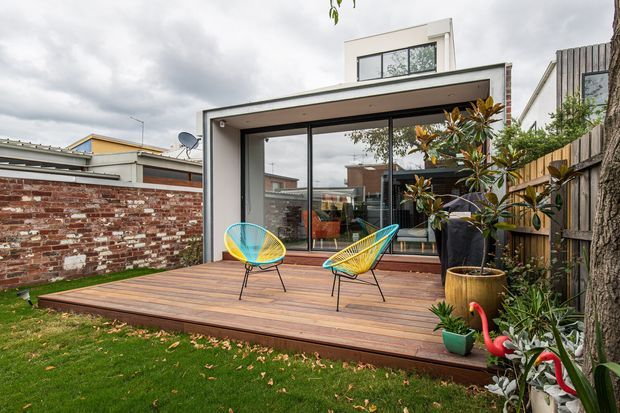 Rear view of a contemporary residential extension with a box-like upper storey, large dark-framed sliding glass doors, and a raised timber deck opening onto a landscaped backyard. The deck is furnished with two colourful wire lounge chairs and potted plants, with neighbouring brick and timber boundary walls visible.