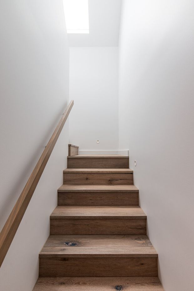 A narrow, minimalist stairway with solid natural timber treads and risers, white painted walls, a simple matching timber handrail, and a small skylight illuminating the upper landing.