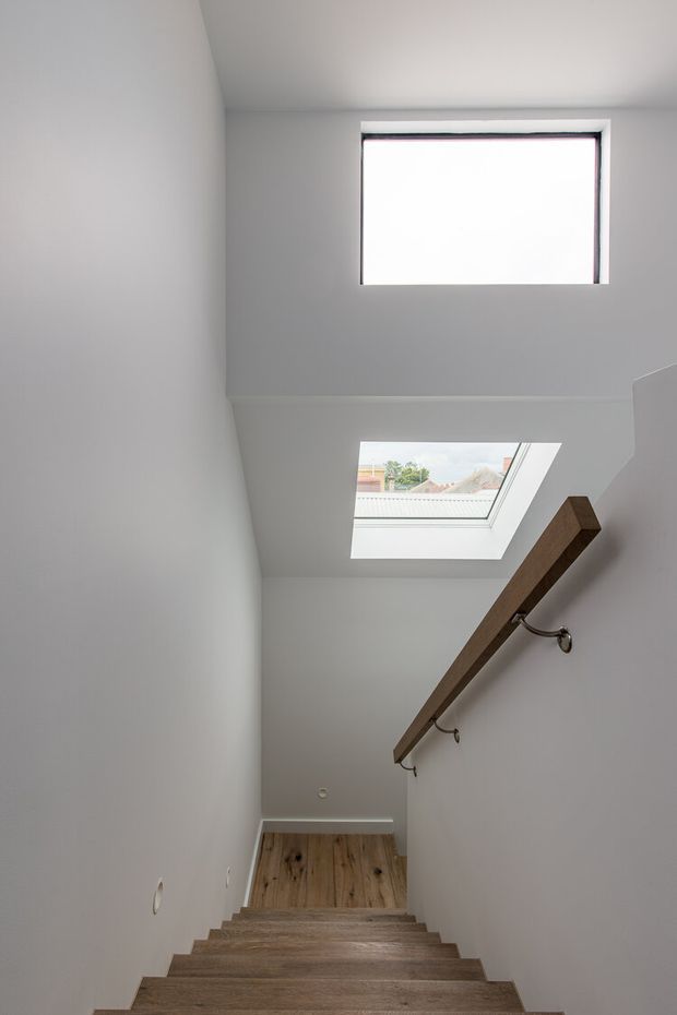A minimalist stairway viewed from below, with white plaster walls and ceiling, warm timber stair treads, and a simple timber handrail supported by dark metal brackets. Two roof windows/skylights bring in natural light, including a rectangular clerestory-style opening above the stairwell.